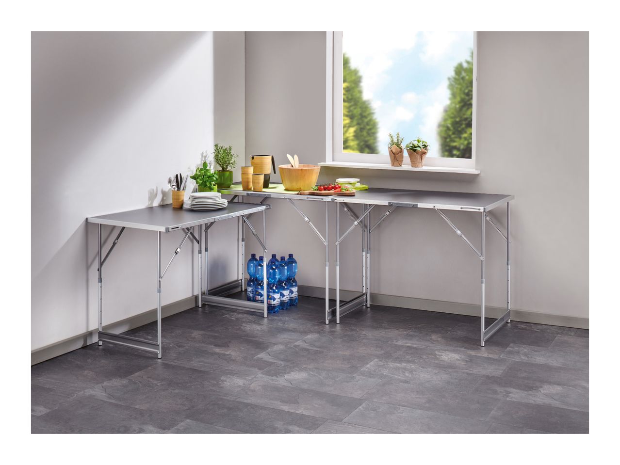 Corner kitchen setup with two folding tables, kitchenware, plants, and bottled water.