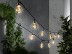 Струна с LED крушки с форма на електрическа крушка, окачена на стена, сред зелени растения.