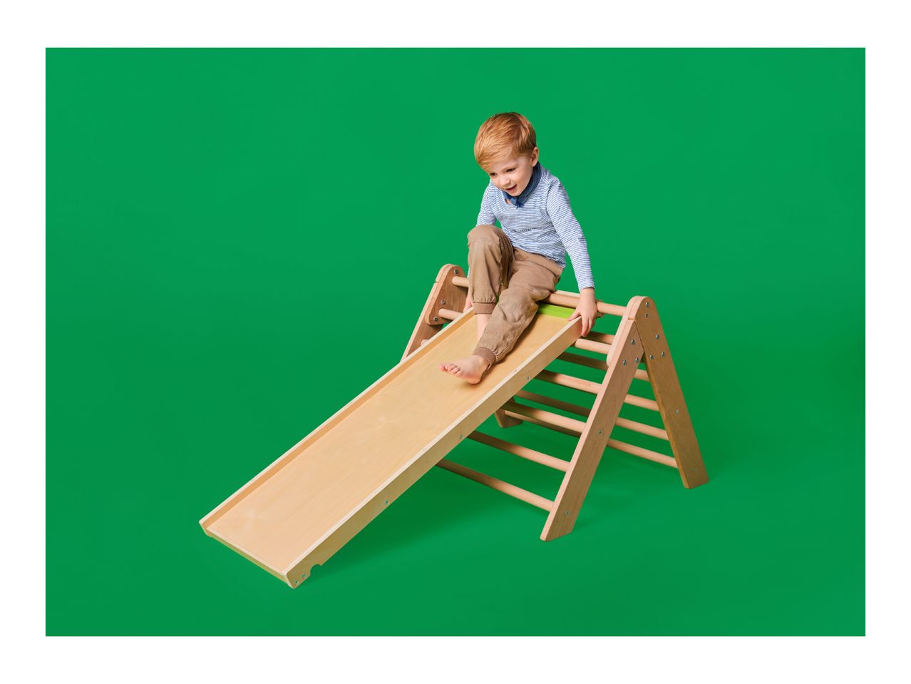 A young boy playing on a wooden indoor slide and climbing frame against a green background.
