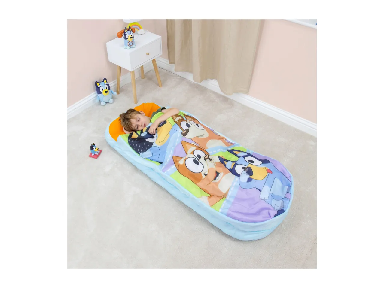 Child sleeping in a colorful character sleeping bag on a carpeted floor with toys nearby.