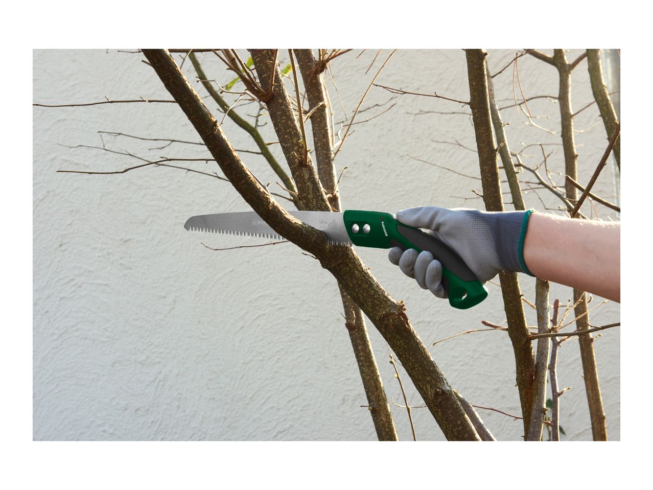 Hand in a grey glove holding a green pruning saw, cutting a tree branch.