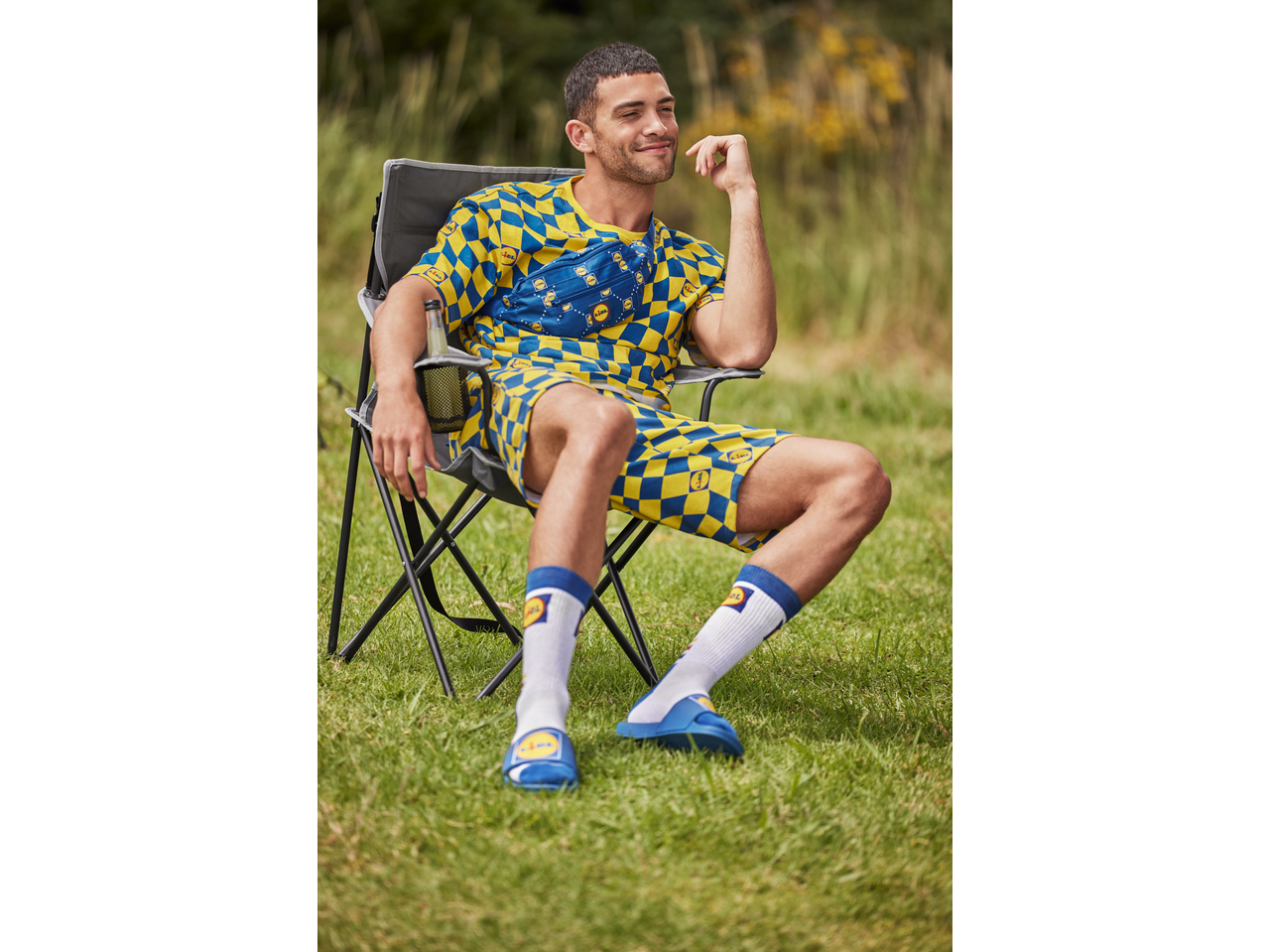 Man in yellow and blue checkered t-shirt, shorts, socks, and slides, sitting in a camping chair.