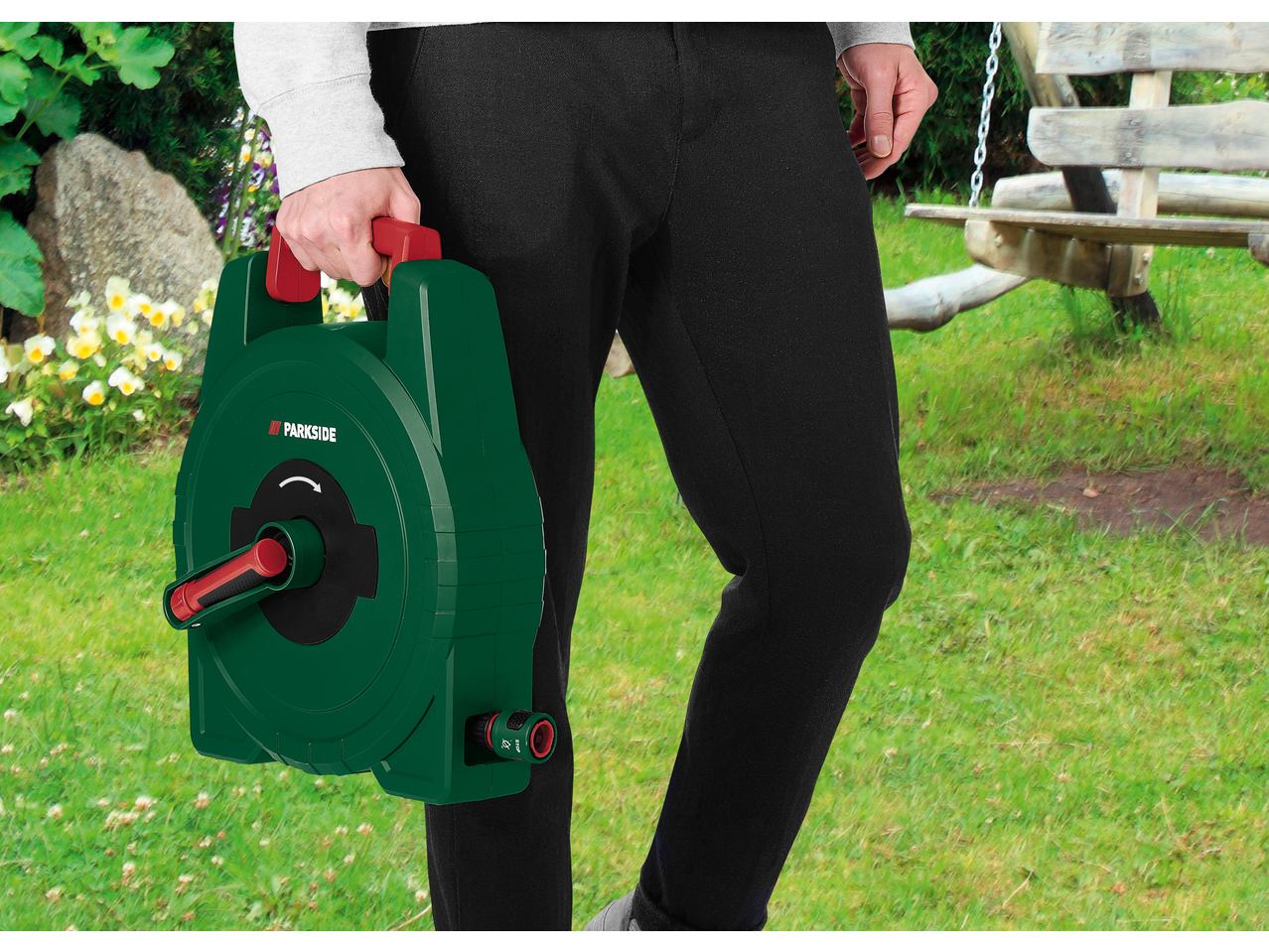 Muškarac sa zelenim koturom za baštensko crevo sa crvenom ručkom, šeta kroz baštu.