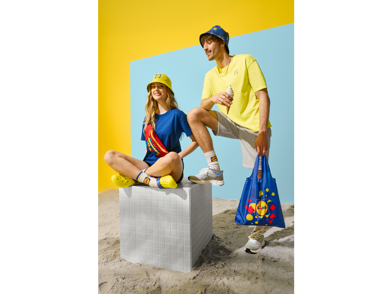 Two models wearing Lidl-branded clothing, including a bum bag, bucket hats, and socks, on a sandy set.