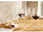 Red wine glasses, some filled, with cheese, grapes, and breadsticks on a wooden table.