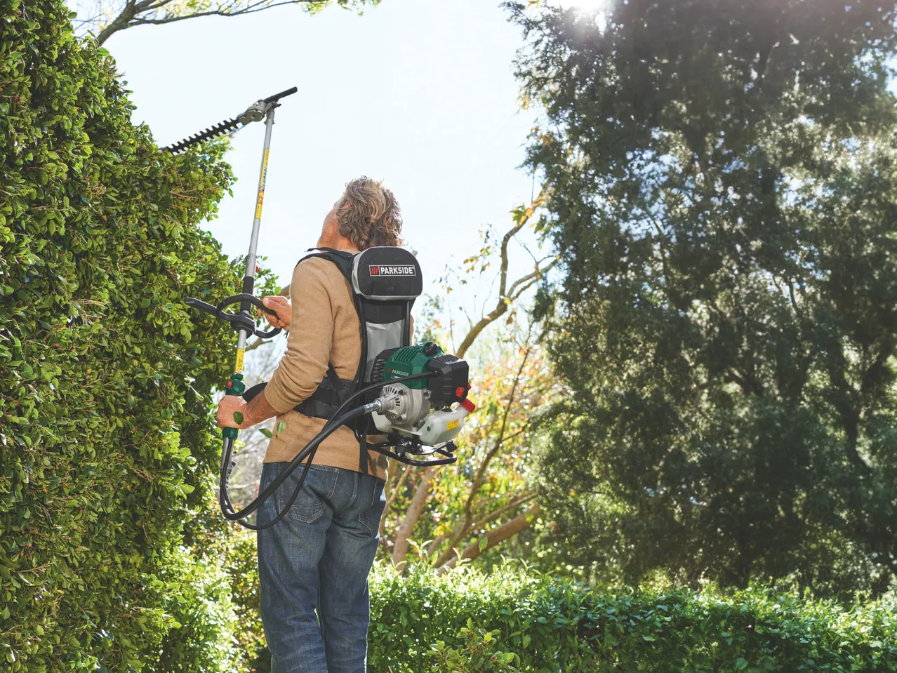 Man trimming a tall hedge with a Parkside Petrol Backpack 6-In-1 Multi Tool with a backpack engine.