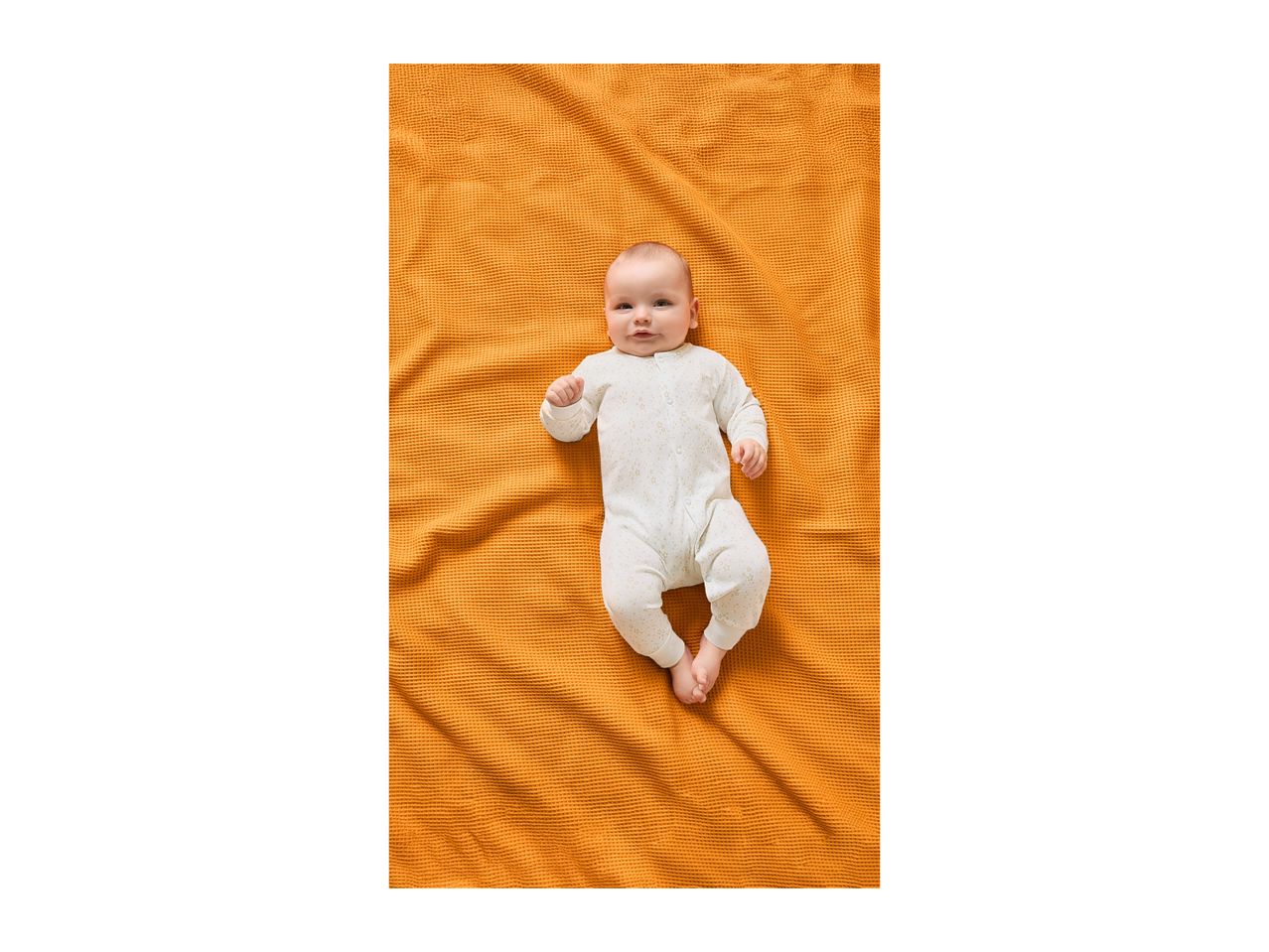 Baby in a white sleepsuit with stars, lying on an orange waffle-knit blanket.