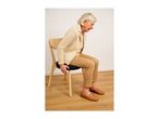 Elderly woman sitting on a chair with a black cushion, wearing a beige cardigan and brown shoes.