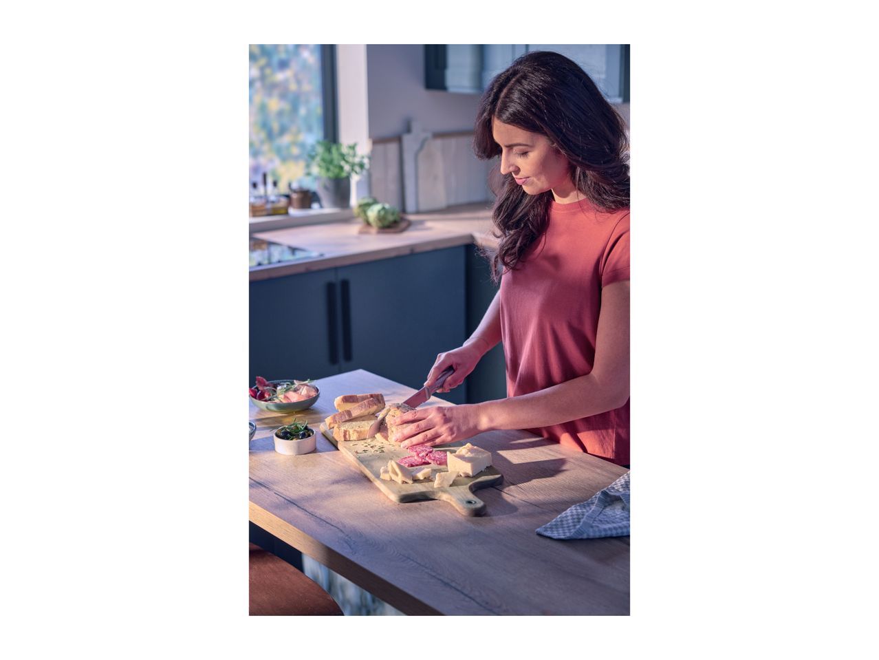 Woman slicing bread on a wooden board with cheese and salami in a modern kitchen.