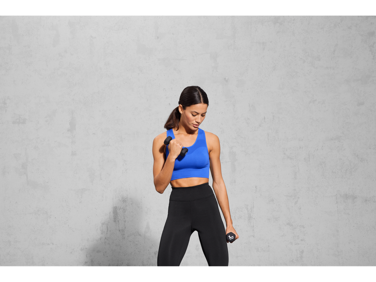 Woman in blue sports bra and black leggings holding two dumbbells.