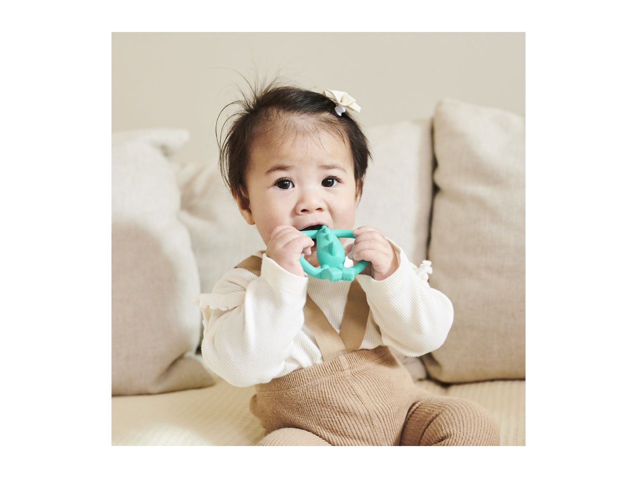 Baby in a white top and brown overalls, chewing on a green teether.