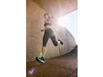 Woman in black sports bra and capri leggings running outdoors, sun flare in background.