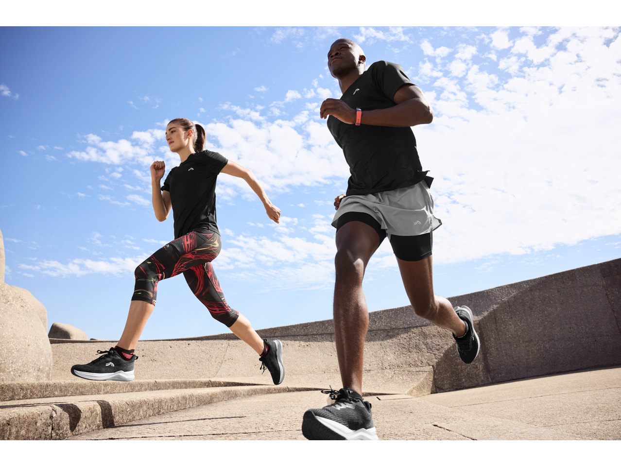 Two runners in black activewear and running shoes, exercising outdoors under a blue sky.
