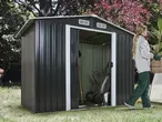 Dark grey garden shed with open door, garden tools inside, and a person carrying a plant.
