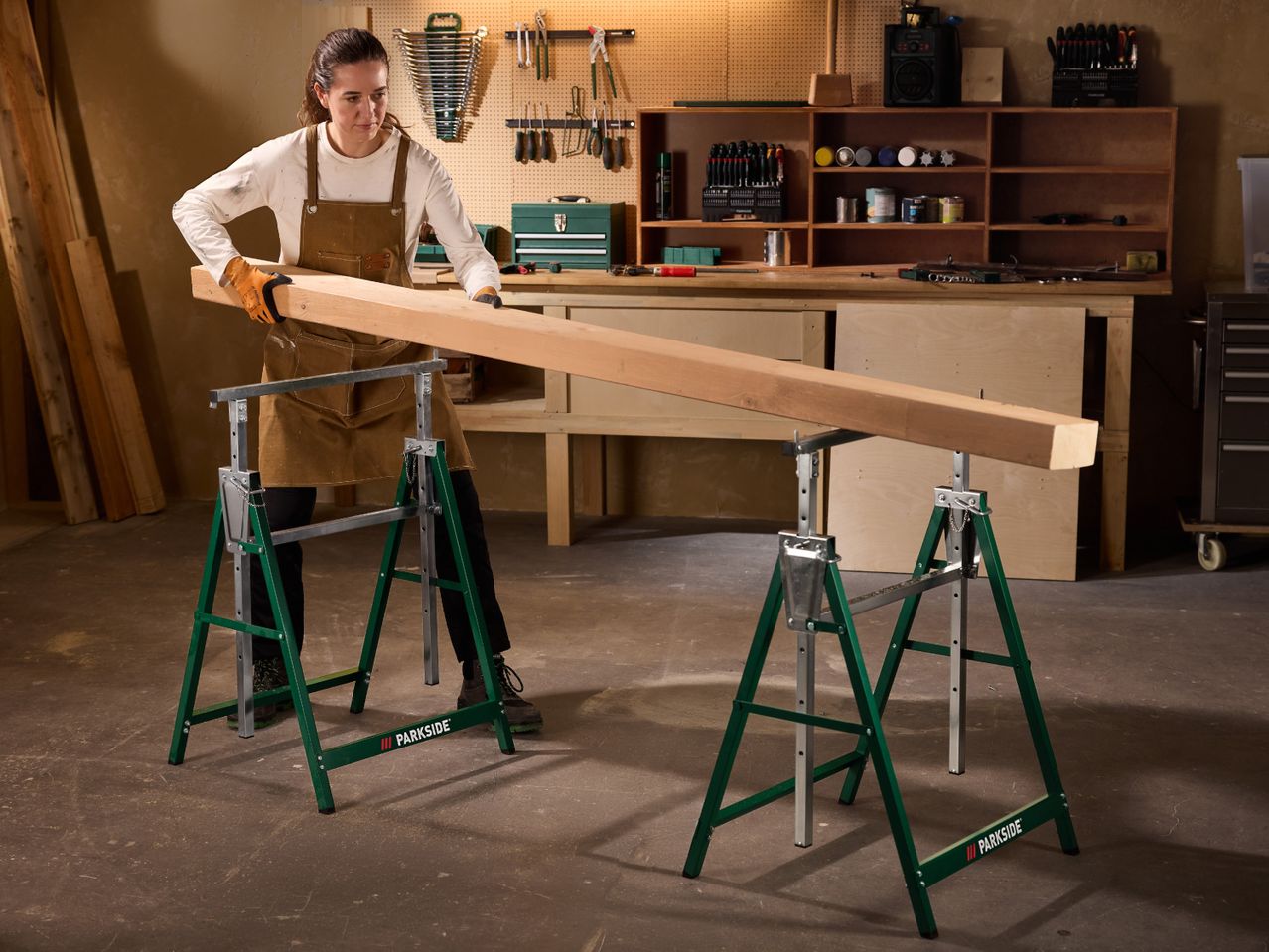 Woman in a workshop placing a wooden beam on two green saw horses.