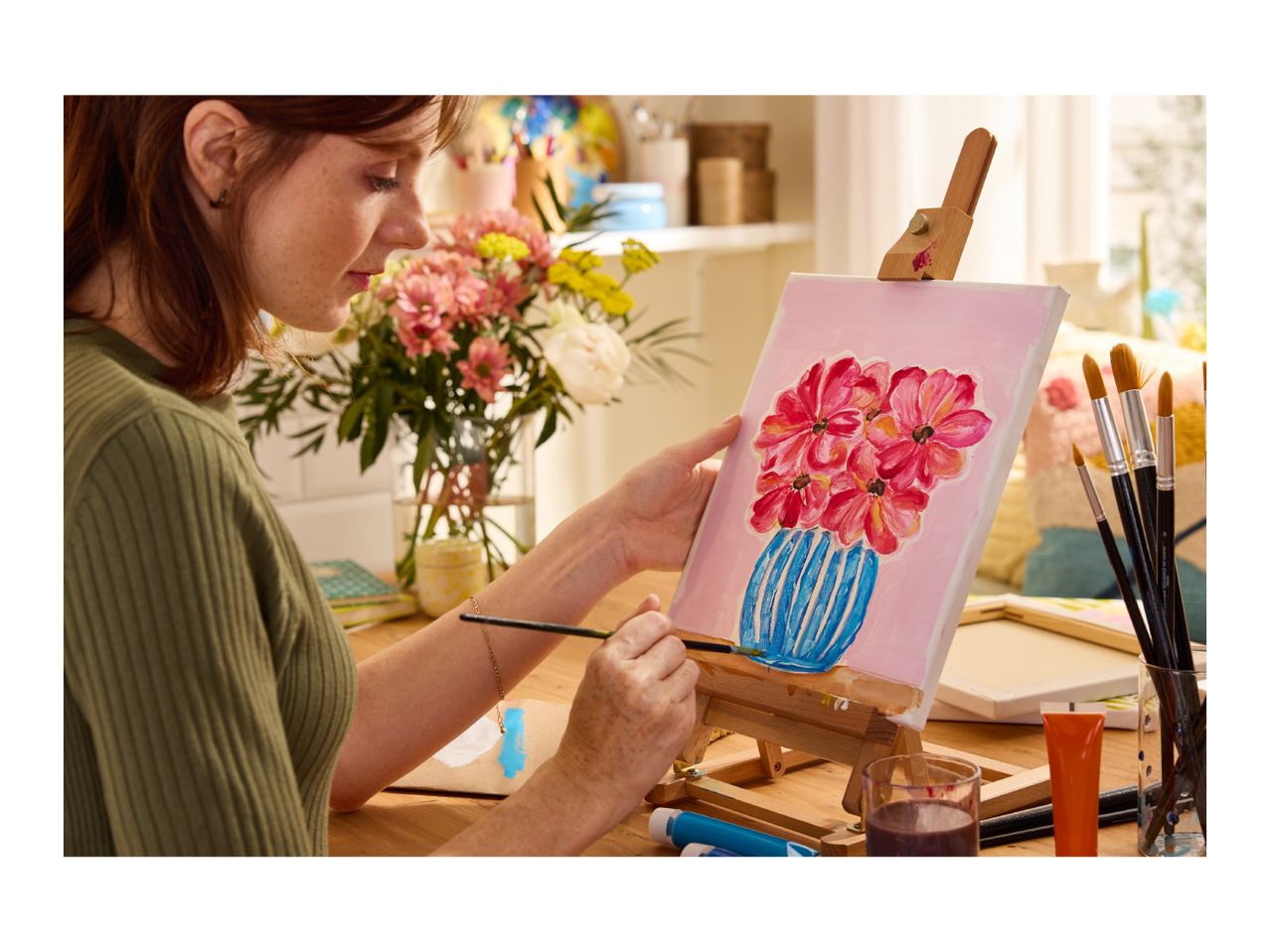Woman painting flowers in a vase on a canvas, surrounded by art supplies and fresh flowers.