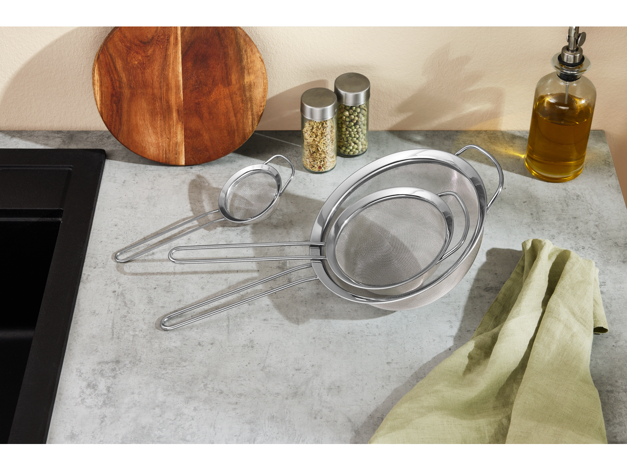 Three stainless steel strainers on a kitchen counter with a wooden board, spices, and olive oil.