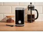 Moedor de café preto, recipiente com café moído e prensa francesa em superfície de madeira.