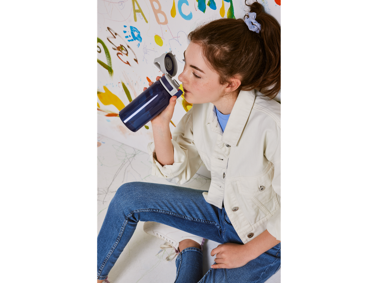 A girl sits on the floor, drinking from a Contigo water bottle against a colorful wall.
