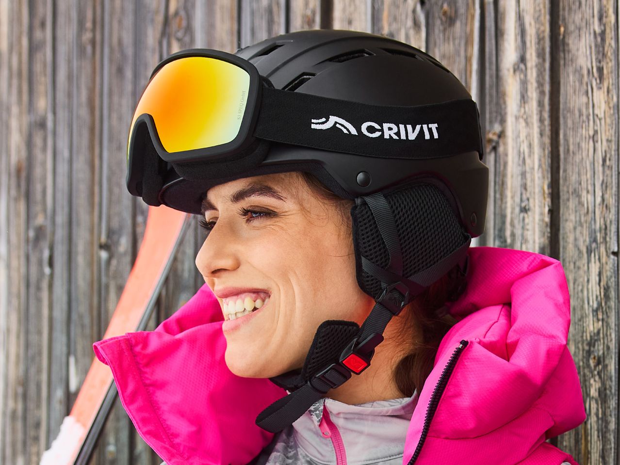 Woman in a black CRIVIT ski helmet and goggles, wearing a bright pink ski jacket.