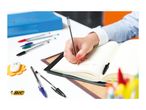 Hand writing in a notebook with a pen, surrounded by other pens and office supplies.