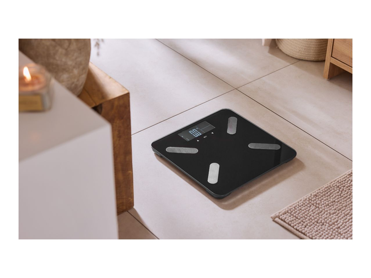 Black smart bathroom scale on a tiled floor, next to a lit candle and wooden furniture.