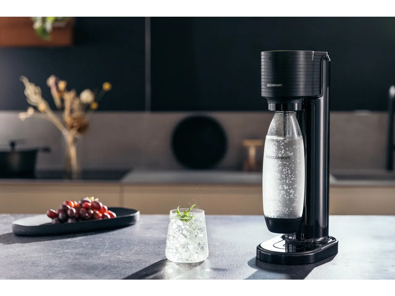 Szódagép, pohár itallal és szőlő egy konyhapulton.