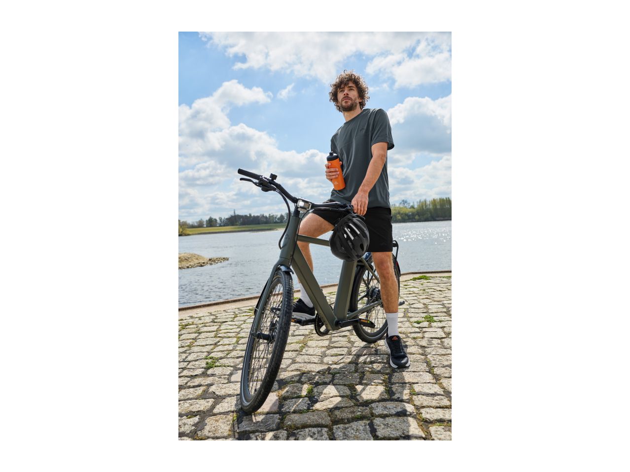 Man with curly hair holding a water bottle, standing next to a bicycle on a cobblestone path.