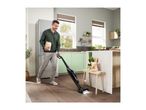 Man vacuuming kitchen floor with a cordless stick vacuum cleaner, holding a bag of snacks.