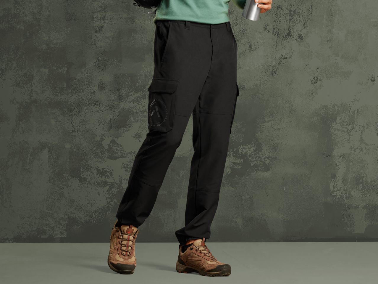 A person wearing black cargo pants and brown hiking boots, holding a silver bottle.
