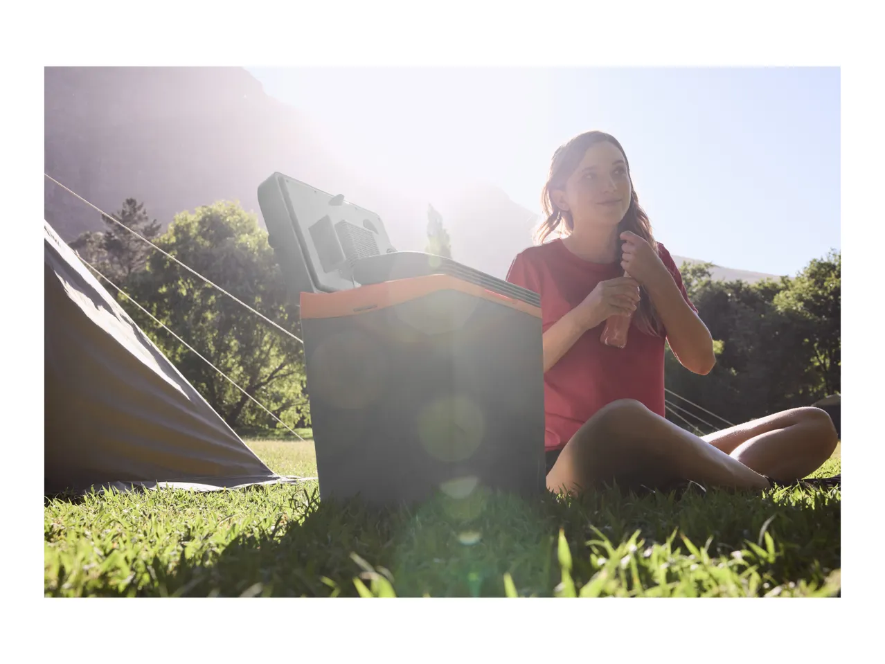 Woman enjoying a drink next to a portable cooler and tent at a campsite.