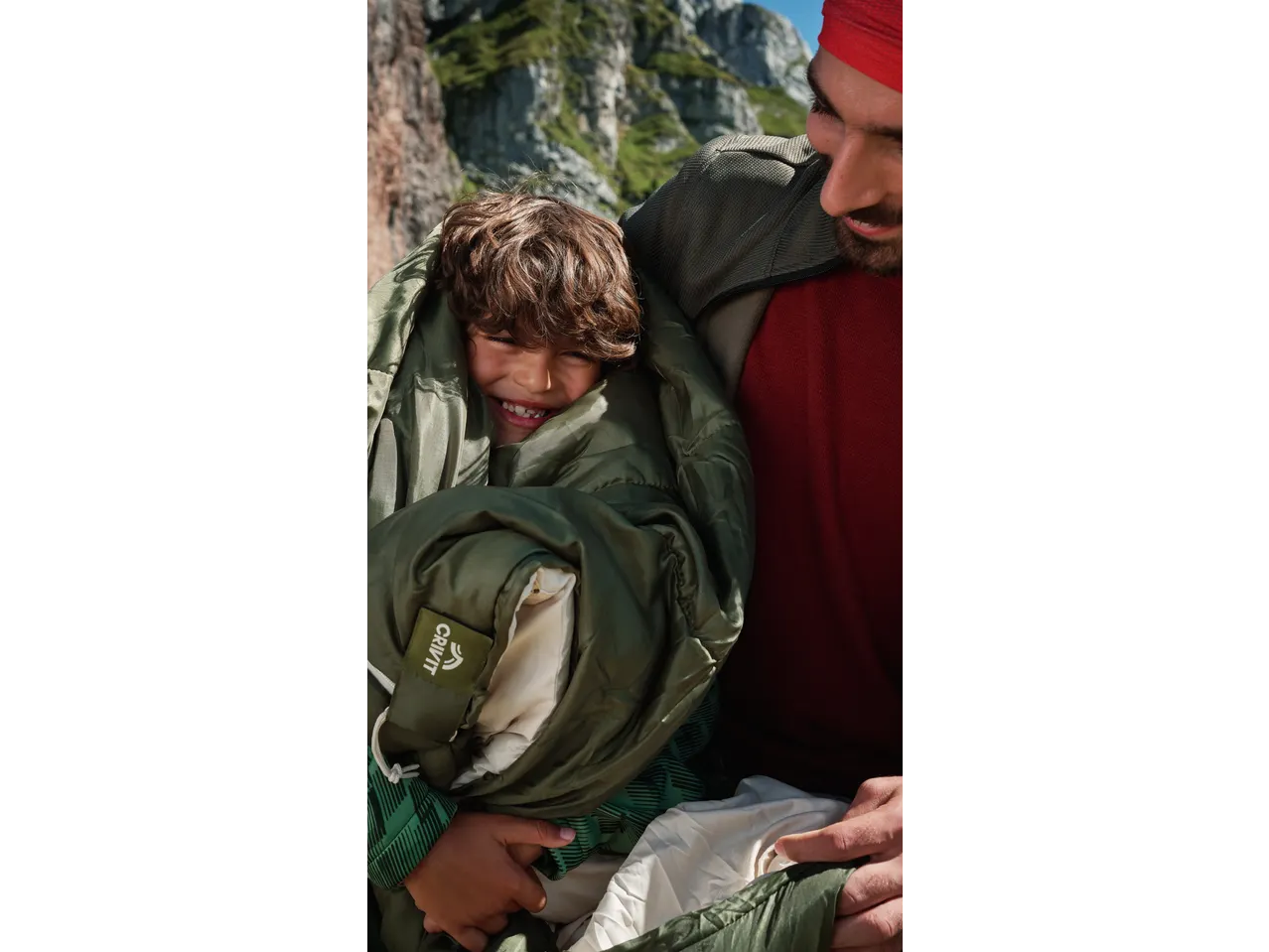 Happy child in a CRIVIT Mummy Sleeping Bag, held by an adult outdoors.