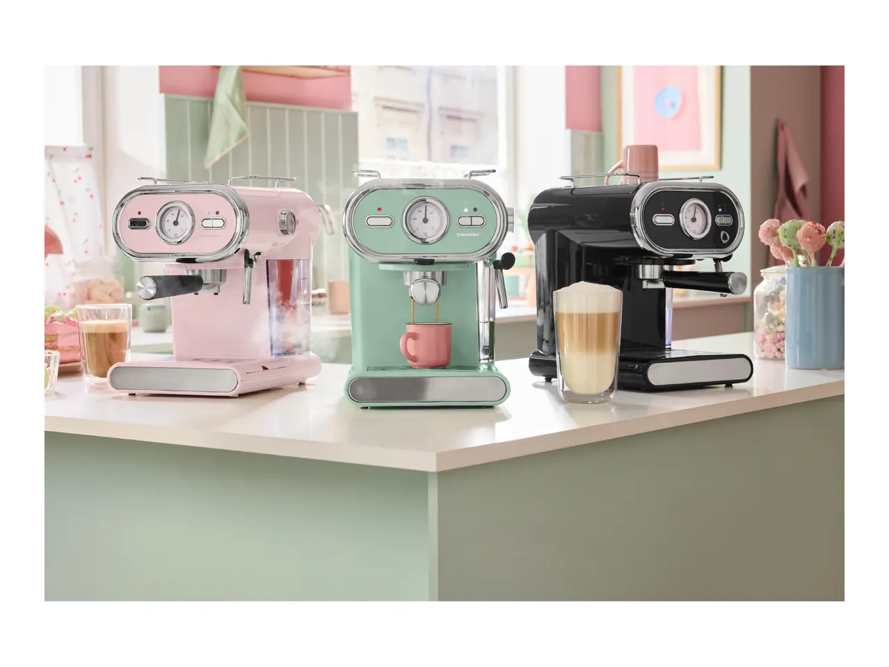 Three retro-style espresso machines in pink, mint green, and black on a kitchen counter.