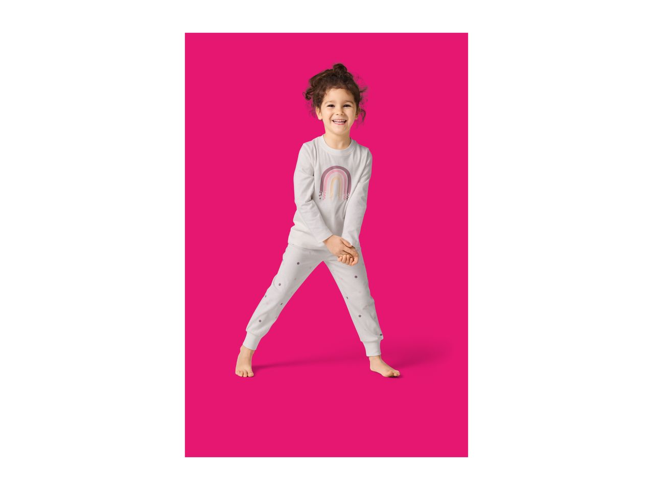 Smiling girl in white pajamas with a rainbow print, standing on a pink background.