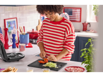 Woman preparing a sandwich with tomato, lettuce, and ham, with a sandwich maker beside her.