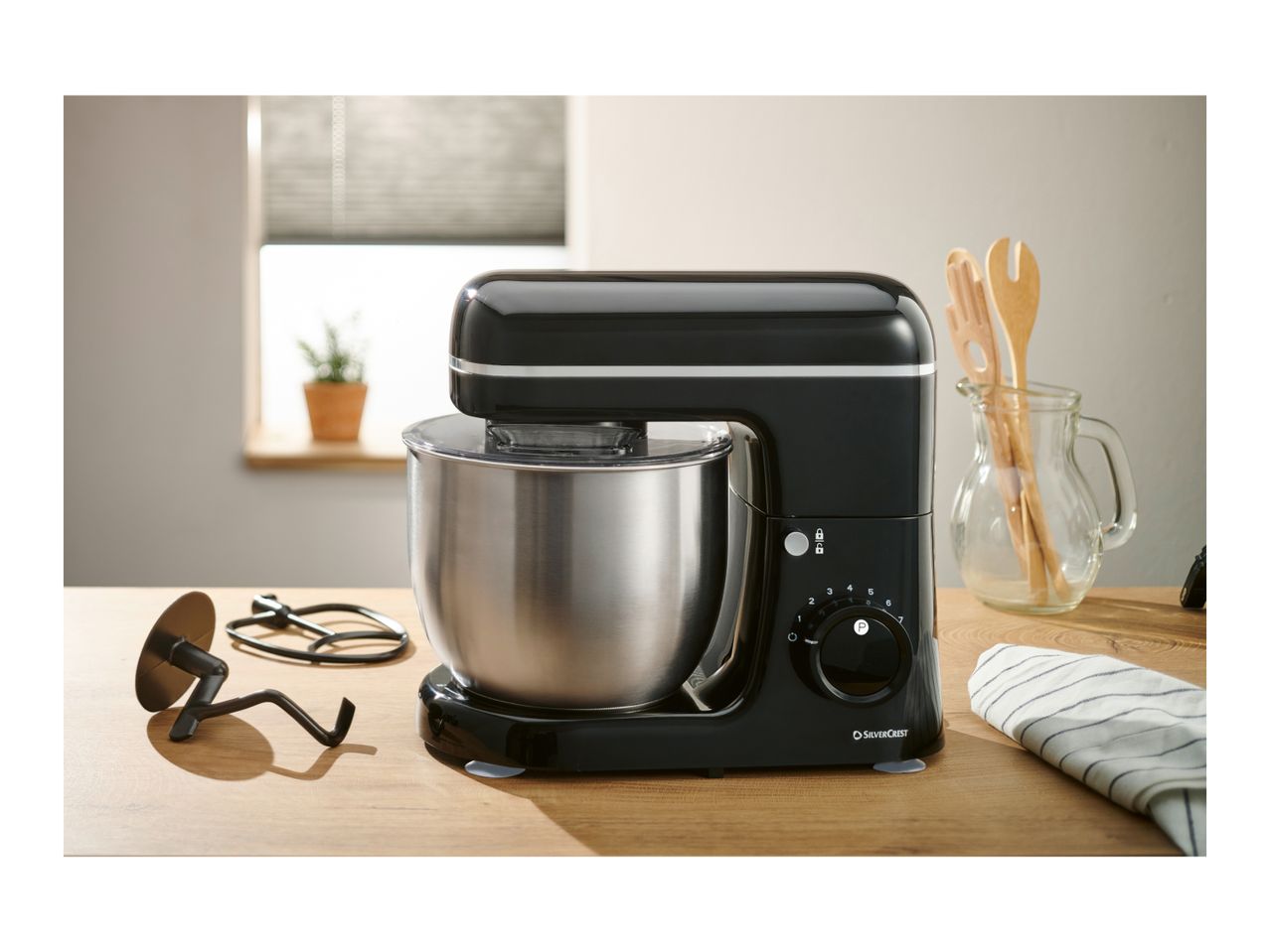 Black stand mixer with a stainless steel bowl on a wooden counter, with kitchen utensils.