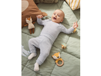 Baby in light blue ribbed sleepsuit, lying on a green blanket with soft toys and wooden rattles.