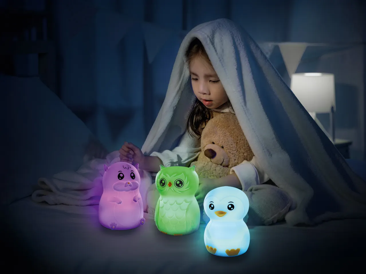 Mädchen mit Decke und Teddybär, umgeben von drei leuchtenden Tier-Nachtlichtern.