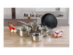 A set of stainless steel pots and pans with glass lids, and a black frying pan, on a kitchen counter with vegetables.