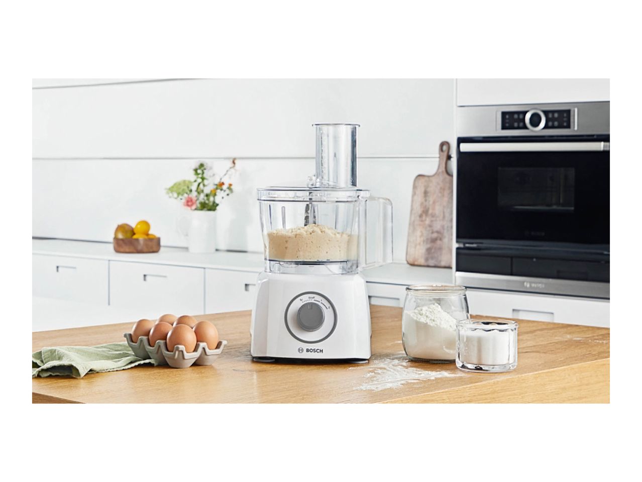 Food processor with dough, eggs, flour, and sugar on a kitchen counter.