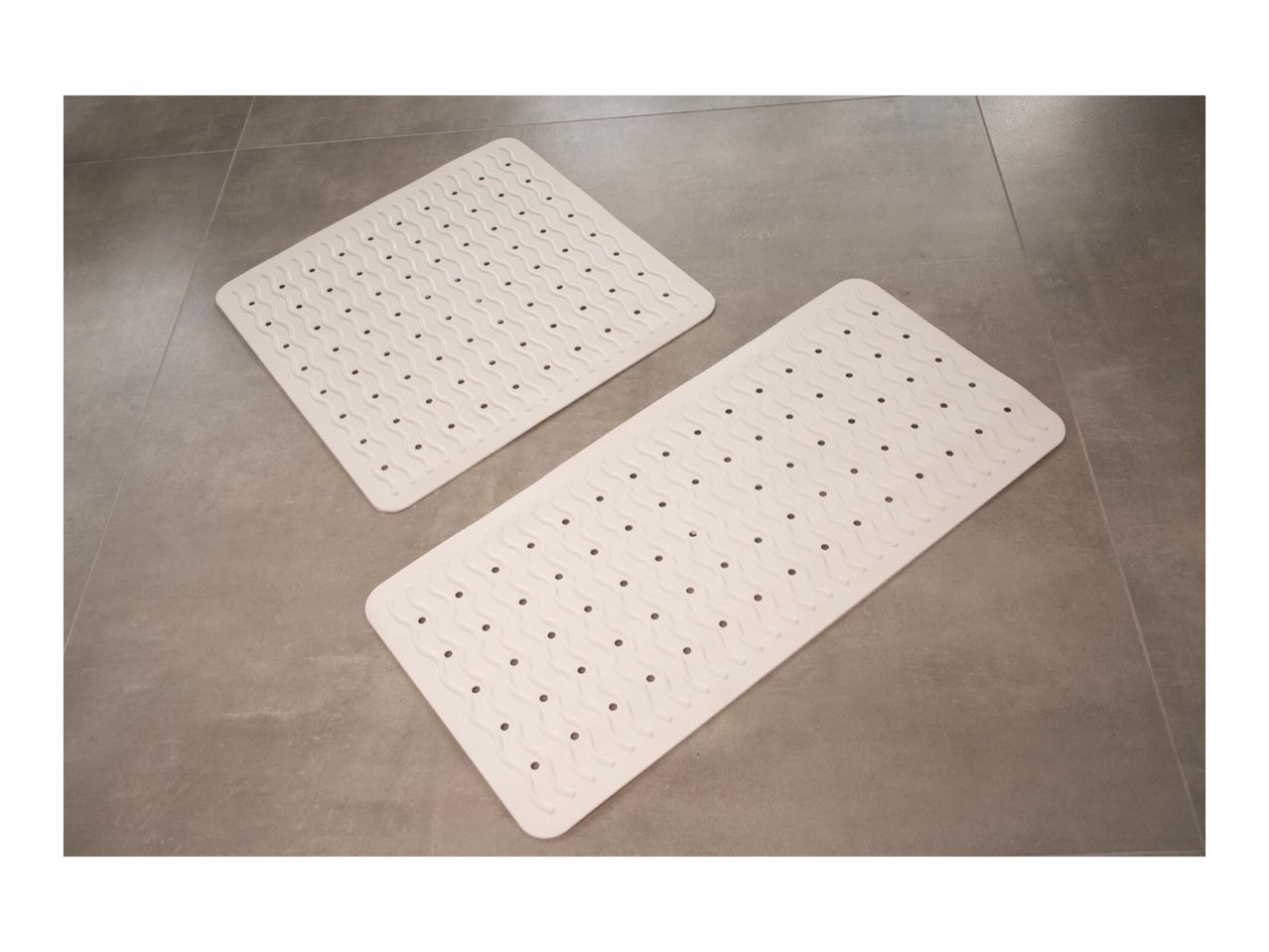 Two rectangular white non-slip bath mats with wavy patterns and drainage holes on a grey tiled floor.