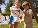 Zwei Kinder hängen Socken und Stofftücher mit Wäscheklammern an eine Wäscheleine im Garten.