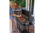 Woman grilling meat and vegetables on a barbecue, with family dining in the background.