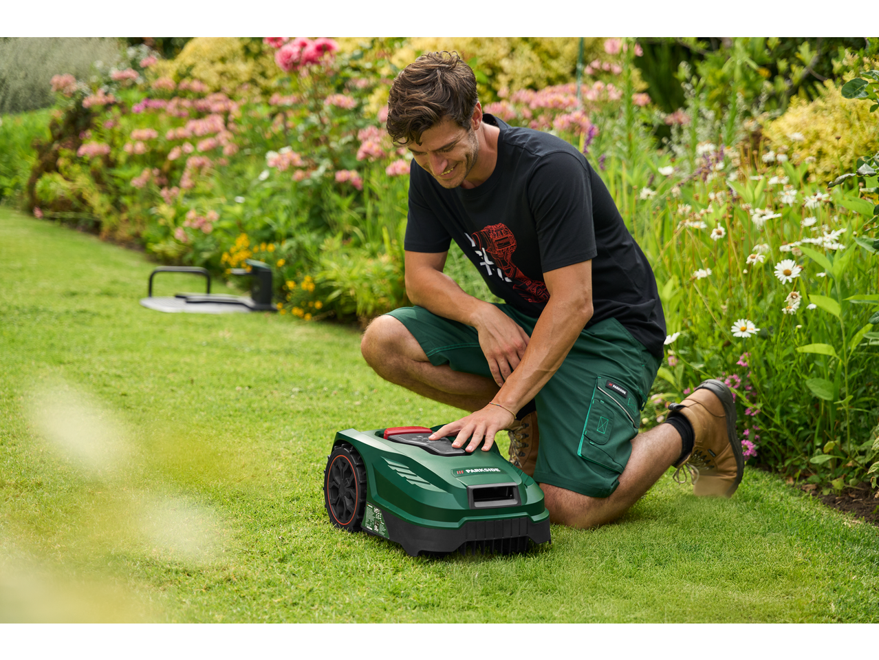 Man setting up a robotic lawnmower in a garden with lush greenery and flowers.