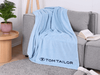 Hellblaue Tom Tailor Fleecedecke auf einem grauen Sofa mit einem Buch und einer Pflanze