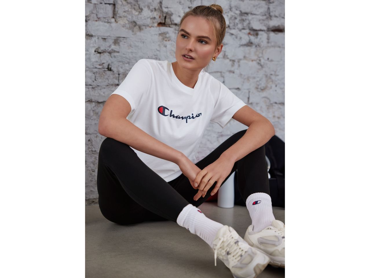 Млада жена, седнала на пода, облечена в бяла тениска Champion и черни клинчета.