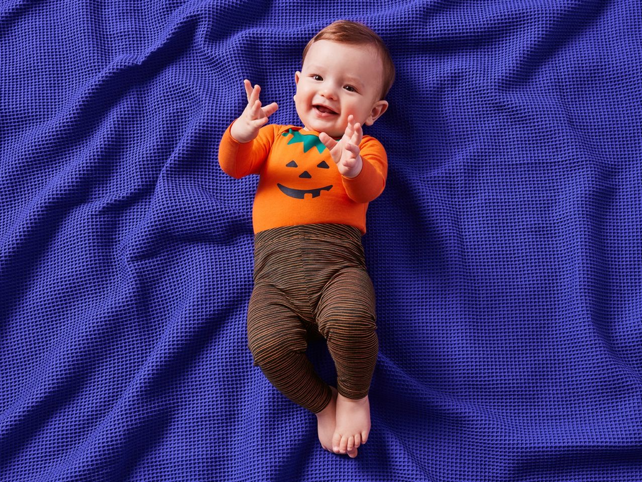 Baby in a pumpkin-themed outfit on a purple waffle-knit blanket.