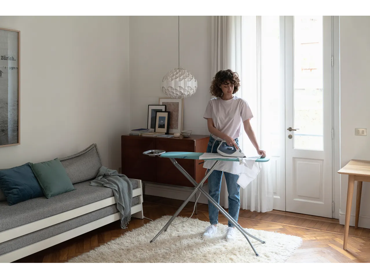 Femeie călcând o cămașă albă pe o masă de călcat într-un living luminos.