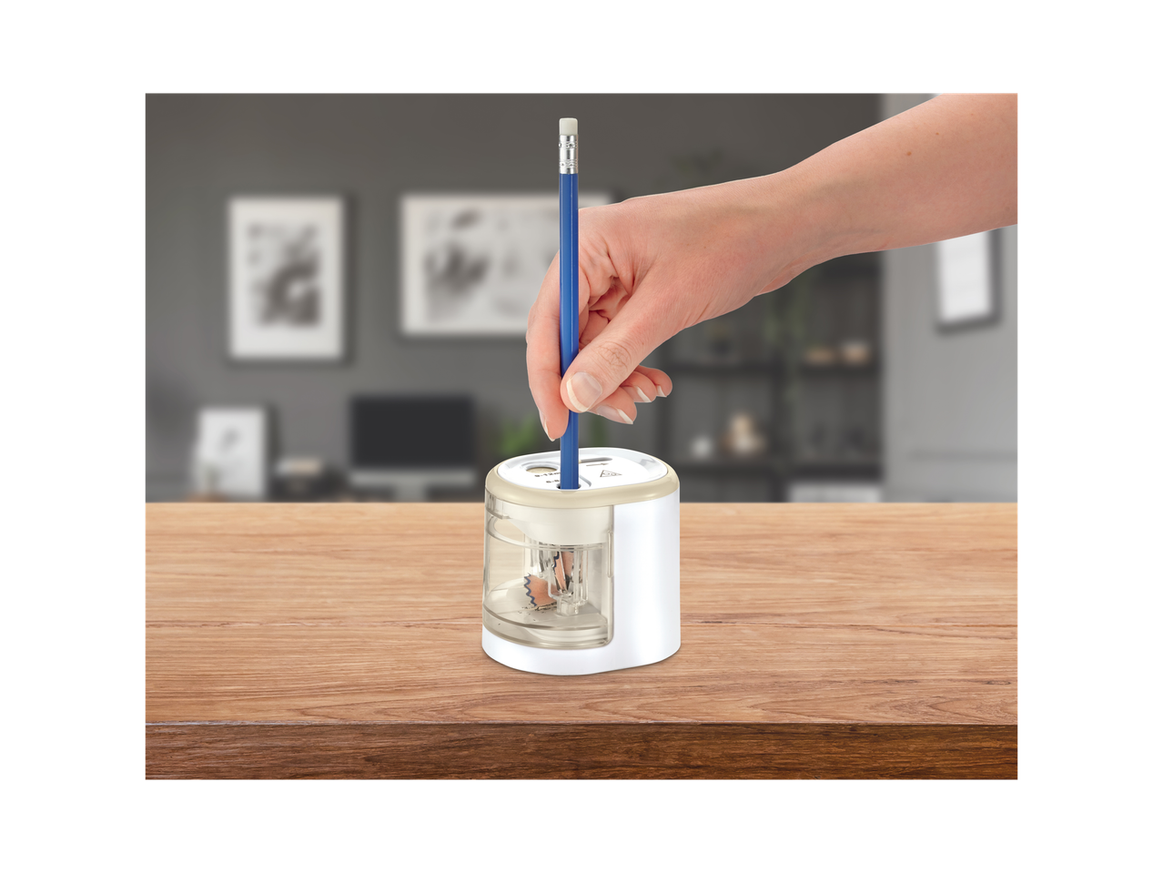 A hand inserts a blue pencil into a white and clear electric pencil sharpener on a wooden table.