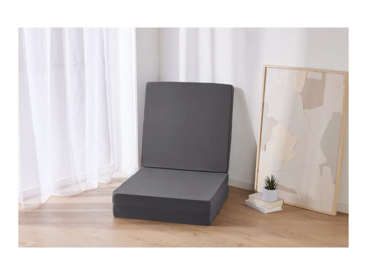 A gray floor chair in a room with white curtains and a framed abstract art piece.